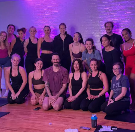 Cheerful group of adults in workout clothes posing for a post-class photo in a purple-lit yoga/fitness studio with exposed brick wall, hardwood floor, mats and water bottles.