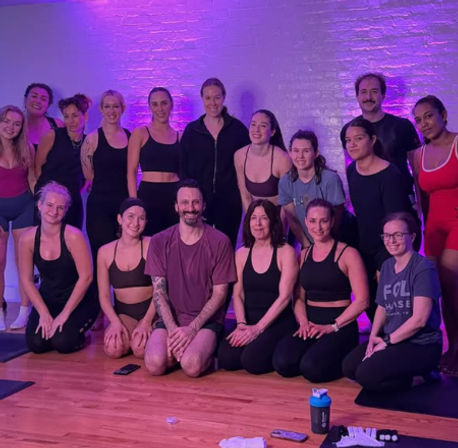 Cheerful group of adults in workout clothes posing for a post-class photo in a purple-lit yoga/fitness studio with exposed brick wall, hardwood floor, mats and water bottles.