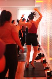 Energetic group fitness class in a narrow brick-wall studio, participants barefoot on mats dancing and stretching under warm red lighting.