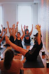Energetic group yoga class in a bright brick-walled studio practicing boat pose, participants balancing on mats with legs lifted and arms reaching up.