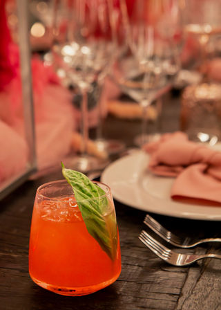 Vibrant orange cocktail with a fresh basil leaf in a short glass on a dark wooden table, beside a white plate, pink napkin and forks with blurred wine glasses in the background.