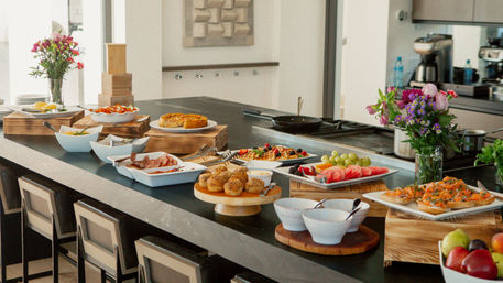 Sunlit modern kitchen island set for brunch with waffles, muffins, pastries, sliced fruit, smoked salmon on toast, bowls and fresh floral arrangements