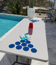 Poolside folding table set up for beer pong with stacked red plastic cups, blue circular targets arranged in triangles and ping-pong balls, on a sunny terrace by a palm tree and rocky hillside