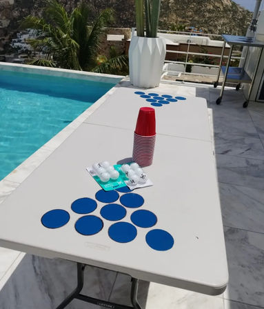 Poolside folding table set up for beer pong with stacked red plastic cups, blue circular targets arranged in triangles and ping-pong balls, on a sunny terrace by a palm tree and rocky hillside