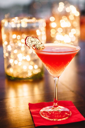 Vibrant pink cocktail in a martini glass with a dragon-fruit slice garnish on a red napkin, warm bokeh string lights over a wooden bar surface.