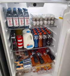Open refrigerator packed with rows of bottled water, stacked egg cartons, a gallon of milk, assorted colored sports drink bottles, cream cheese and deli meats on lower shelves.