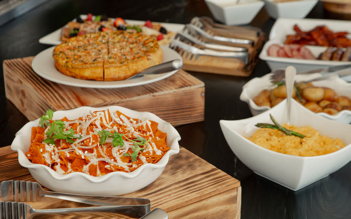 Brunch buffet on dark table with white serving bowls on wooden risers: saucy tortilla-chip chilaquiles topped with crema and cilantro, sliced quiche, scrambled eggs, roasted potatoes and breakfast meats.