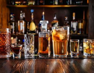 Assorted whiskey glasses, shots and an ice-filled cocktail lined up on a wooden bar counter with blurred liquor bottles on the back bar — a cheers-ready lineup.