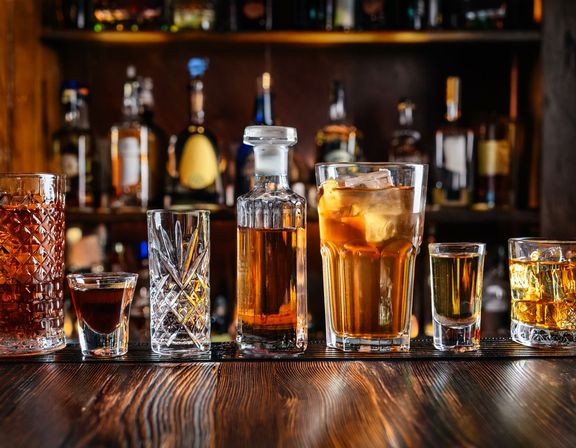Assorted whiskey glasses, shots and an ice-filled cocktail lined up on a wooden bar counter with blurred liquor bottles on the back bar — a cheers-ready lineup.