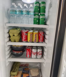 Neatly stocked kitchen refrigerator with bottled water on top, stacks of canned soda, egg cartons, deli meats, cheese and meal-prep bowls on shelves.