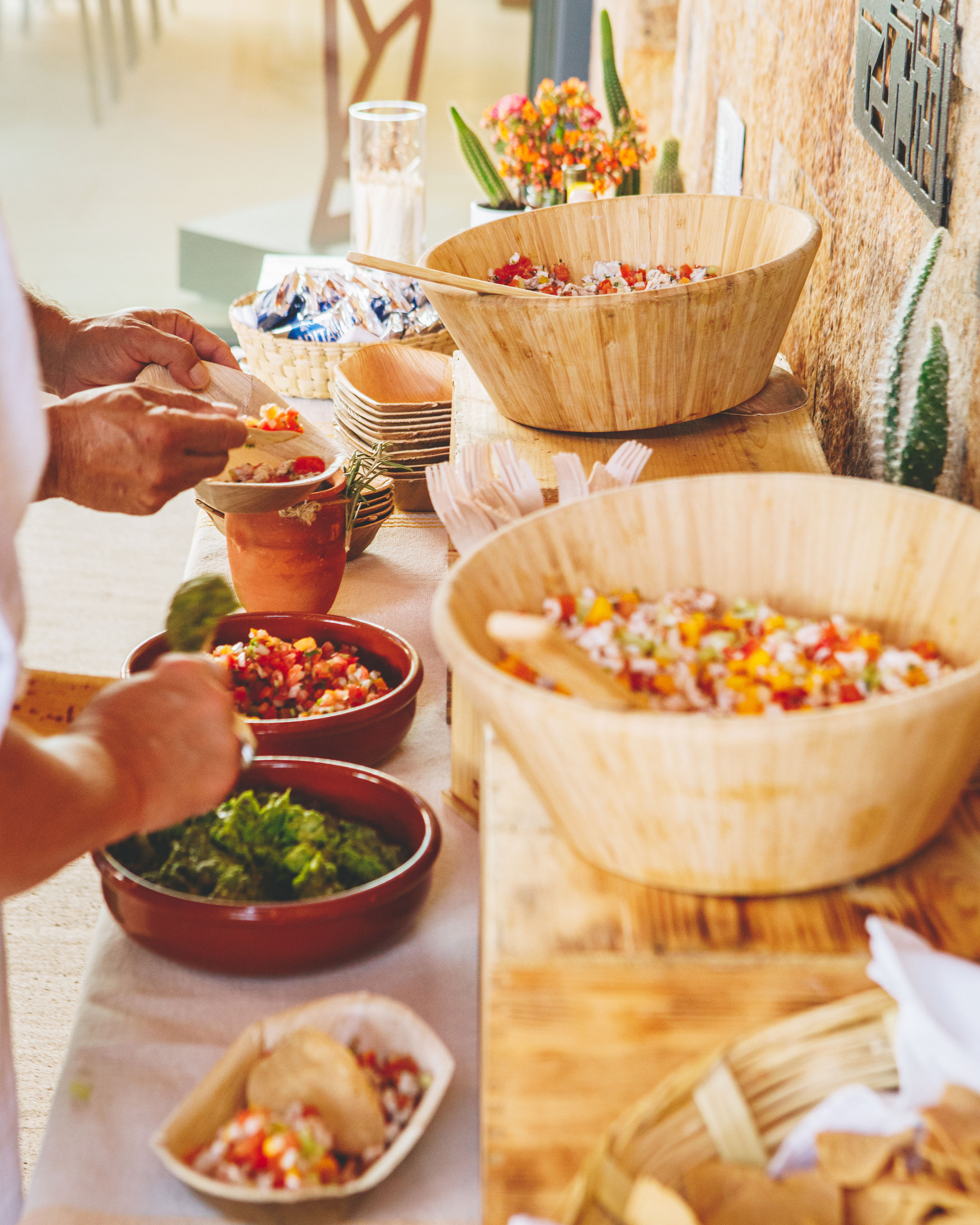 Outdoor buffet table with wooden bowls of colorful salsa and salad, terracotta dishes and taco shells, hands serving toppings beside a small potted cactus