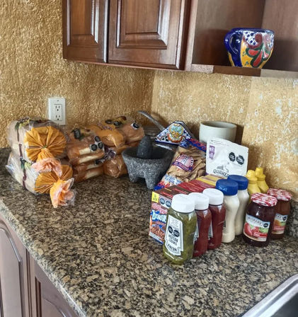 Granite kitchen countertop with stacked packaged buns, a stone molcajete mortar, assorted condiments and jams, cereal packets, and a colorful ceramic mug under wooden cabinets.