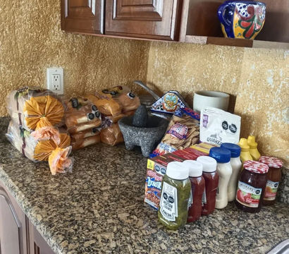 Granite kitchen countertop with stacked packaged buns, a stone molcajete mortar, assorted condiments and jams, cereal packets, and a colorful ceramic mug under wooden cabinets.