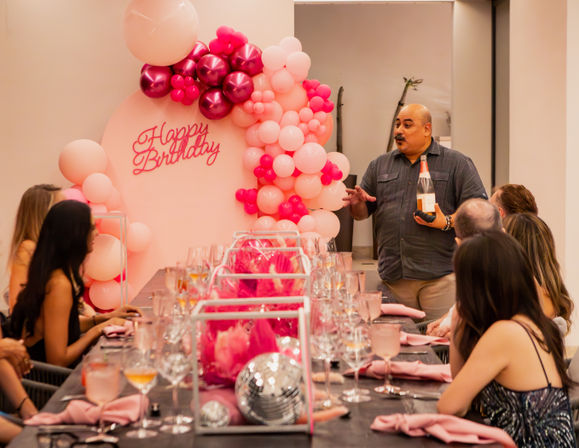 Cheerful indoor birthday dinner in a private event room with a pink balloon arch and “Happy Birthday” sign, long table set with wine glasses, pink napkins and disco-ball decor as a host presents a bottle of sparkling wine to seated guests.