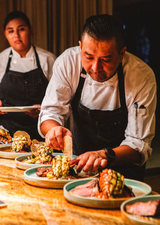 Chef plating gourmet seafood and steak entrees on ceramic plates during dinner service — close-up of hands adding a crisp garnish in a warm-lit restaurant kitchen