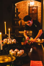 Server arranging cream-topped pastries on a candlelit dessert table in a warm brick courtyard, with lit taper candles and small potted succulents.