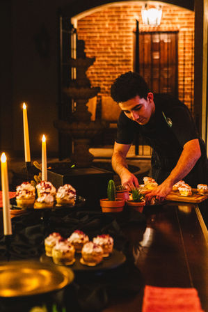 Server arranging cream-topped pastries on a candlelit dessert table in a warm brick courtyard, with lit taper candles and small potted succulents.