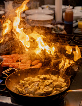 Fiery commercial stovetop with sausages grilling and mushrooms sizzling in a skillet — lively restaurant kitchen action.