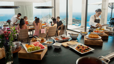 Vibrant ocean-view brunch buffet at a sunlit coastal villa — fresh fruit, waffles, pastries and a chef serving a relaxed group by floor-to-ceiling windows.