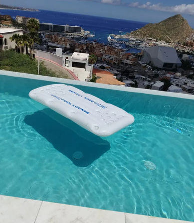 White inflatable pool lounge floating in a crystal-clear infinity pool overlooking a coastal marina, blue ocean and rocky headland under a sunny sky
