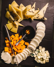 Overhead shot of a cheese board on dark slate: triangular marbled hard-cheese wedges, bright orange cheddar cubes, a curved row of soft white cheese slices, blue cheese cubes, and a small bowl of honey with a spoon.