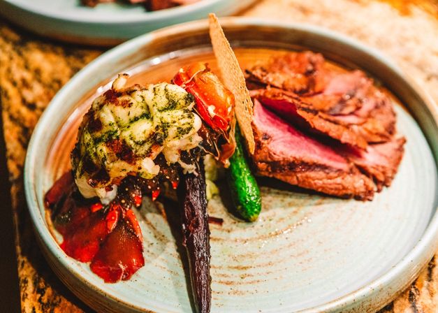 Gourmet surf-and-turf plate with grilled lobster tail topped with herb butter, roasted baby carrot and zucchini, a crisp cracker, and sliced medium-rare beef on a ceramic plate — fine-dining presentation.