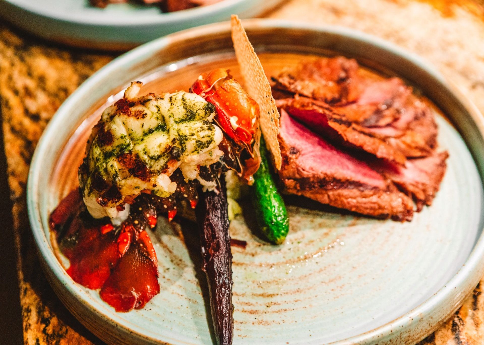 Gourmet surf-and-turf plate with grilled lobster tail topped with herb butter, roasted baby carrot and zucchini, a crisp cracker, and sliced medium-rare beef on a ceramic plate — fine-dining presentation.
