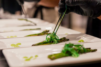 Gloved chef plating grilled asparagus topped with vibrant seaweed salad using tweezers on white rectangular plates in a restaurant kitchen — elegant fine-dining appetizer.