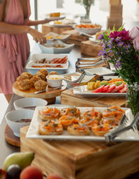 Bright brunch buffet spread at a daytime event with smoked salmon canapés, mini muffins, fresh fruit platter, assorted bowls and a floral centerpiece