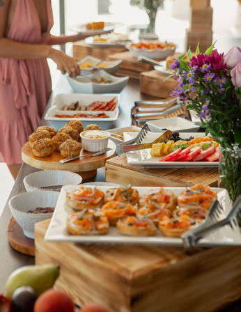 Bright brunch buffet spread at a daytime event with smoked salmon canapés, mini muffins, fresh fruit platter, assorted bowls and a floral centerpiece