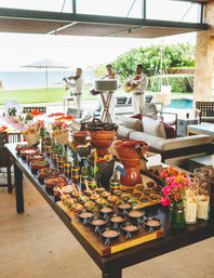 Beachfront patio outdoor buffet with colorful Mexican-style bowls, salsas and tortillas on a long table, clay pots and flowers in the foreground, and a trio of musicians playing near a pool and ocean view.