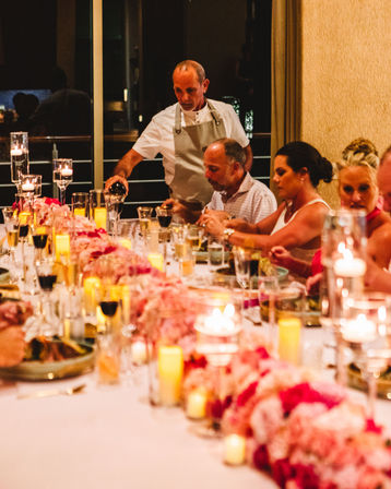 Elegant candlelit evening dinner with guests seated at a long table lined with pink floral centerpieces and glowing votive candles, a server in an apron pouring red wine