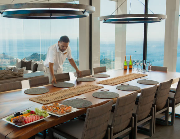 Staff arranging round placemats on a long wooden dining table in a modern coastal dining room with panoramic ocean views, circular pendant lights, champagne bottles and fruit platters.