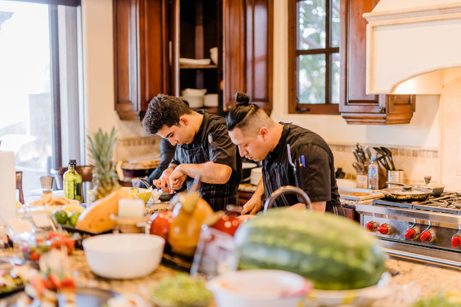 Two chefs in black uniforms prepping food at a busy residential kitchen island with fresh produce, watermelon, pineapple, bowls and a gas range