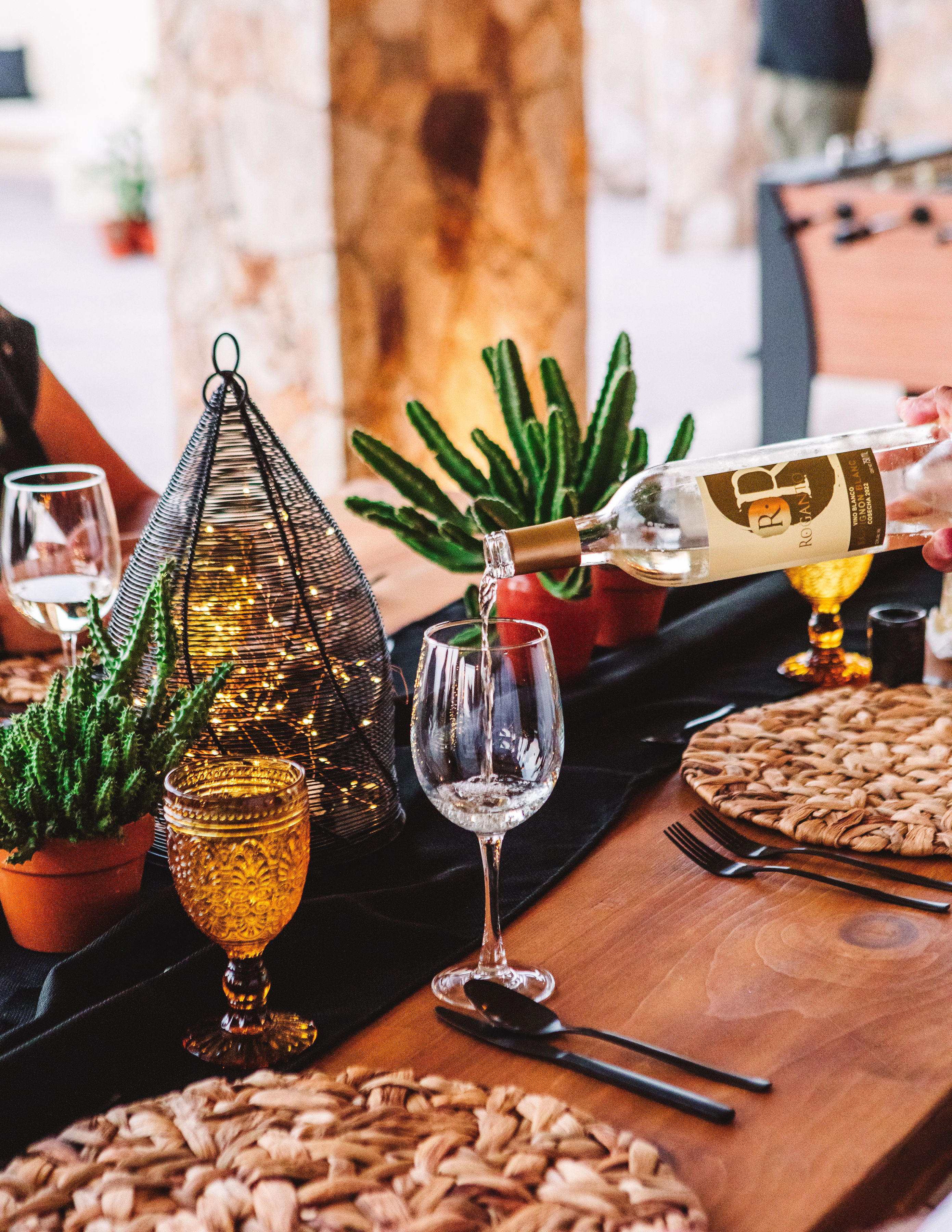 Rustic wooden dinner table with white wine being poured into a glass, amber goblets, woven placemats, potted succulents, and a wire lantern filled with fairy lights.