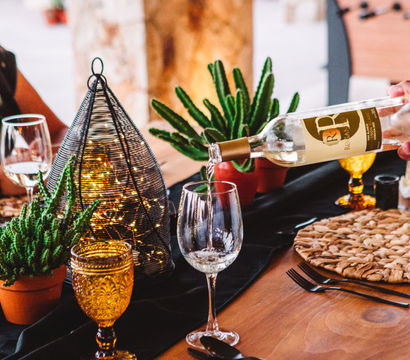 Rustic wooden dinner table with white wine being poured into a glass, amber goblets, woven placemats, potted succulents, and a wire lantern filled with fairy lights.