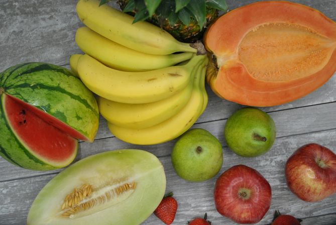 Vibrant assortment of fresh summer fruits—bananas, halved papaya, watermelon, honeydew, pineapple top, green pears, red apples and strawberries—arranged on a weathered gray wooden table.