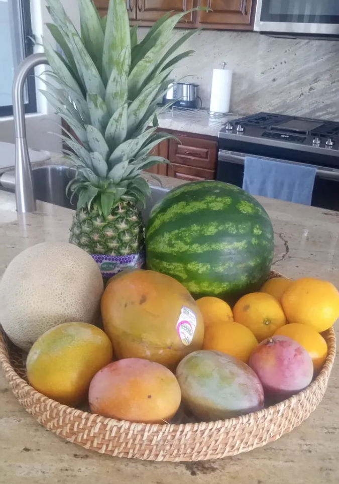 Bright woven fruit basket on a kitchen island with pineapple, whole watermelon, cantaloupe, mangoes and oranges on a modern stone countertop.