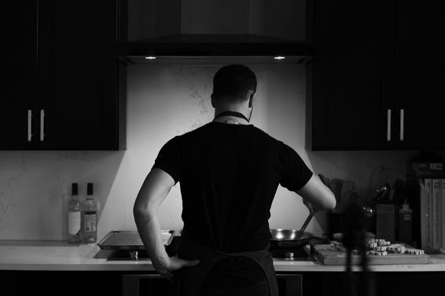 Black-and-white back view of a person in a black shirt and apron stirring a skillet under a range hood in a modern home kitchen, with a cutting board of sliced vegetables and bottles on the countertop.