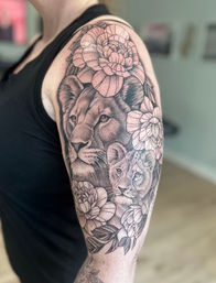 Regal black-and-gray upper arm/shoulder tattoo: realistic lion and cub portraits surrounded by detailed peony flowers and leaves in a half-sleeve design.