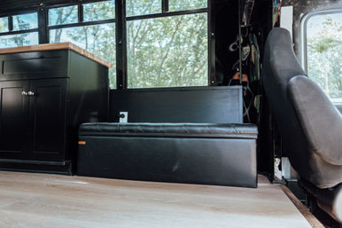 Cozy camper van interior with a black leather storage bench under windows, matching black cabinet with wood countertop, driver's seat on the right, light wood floor and leafy outside view.