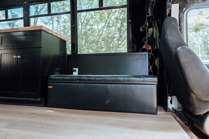 Cozy camper van interior with a black leather storage bench under windows, matching black cabinet with wood countertop, driver's seat on the right, light wood floor and leafy outside view.