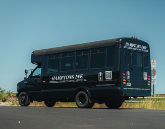 Black mobile tattoo studio bus parked on a coastal road by sand dunes and grasses under a clear blue sky