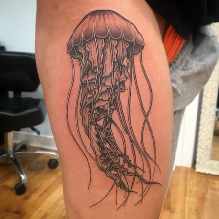 Black-and-grey jellyfish tattoo on upper thigh, detailed linework and shading with long flowing tentacles, photographed in a tattoo studio