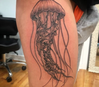 Black-and-grey jellyfish tattoo on upper thigh, detailed linework and shading with long flowing tentacles, photographed in a tattoo studio