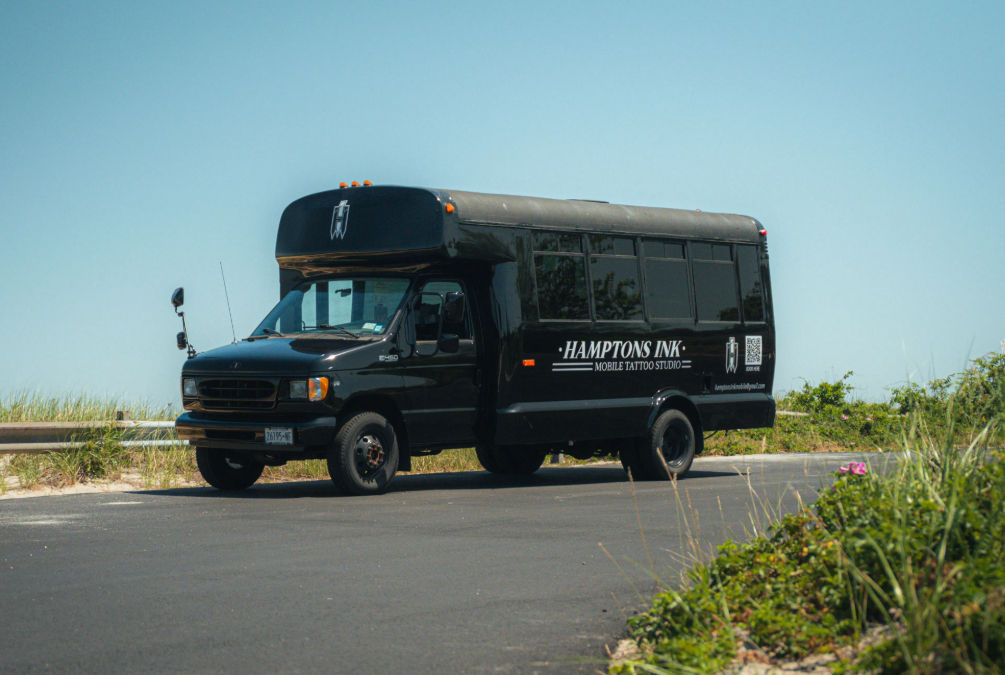 Sleek black converted shuttle bus labeled as a mobile tattoo studio parked on a sunny coastal road beside sand dunes and beach grasses.
