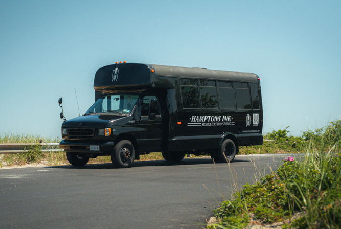 Sleek black converted shuttle bus labeled as a mobile tattoo studio parked on a sunny coastal road beside sand dunes and beach grasses.