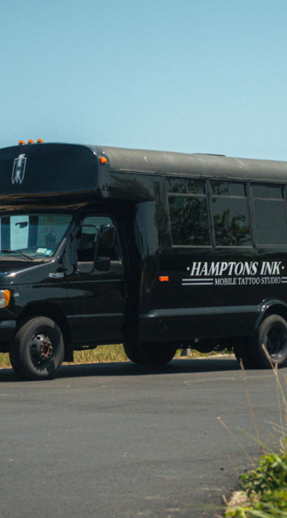Sleek black converted shuttle bus labeled as a mobile tattoo studio parked on a sunny coastal road beside sand dunes and beach grasses.