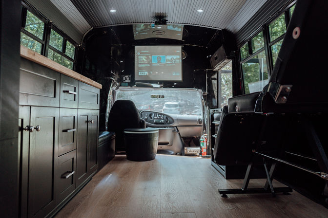 Cozy converted bus interior: wood floor, dark built-in cabinets, projector screen above the driver’s cockpit, seating and large windows showing trees outside.