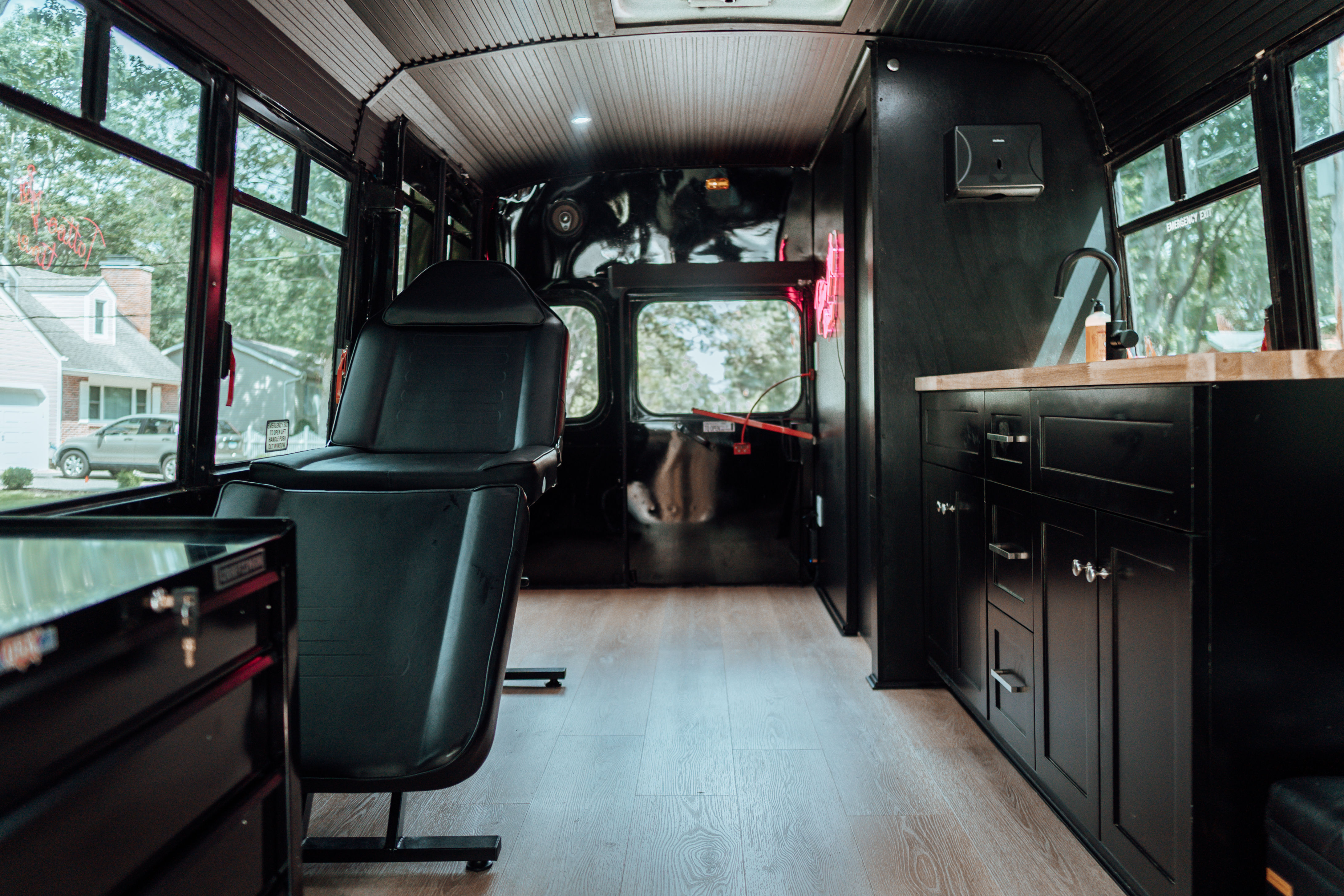 Cozy converted bus mobile studio interior with a black reclining chair, wood floors, matte black cabinets and sink, and large windows overlooking a suburban street.